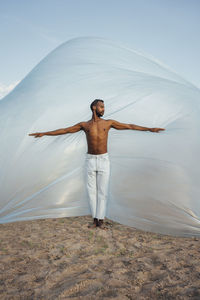 Shirtless man with arms outstretched standing in front of foil on sand