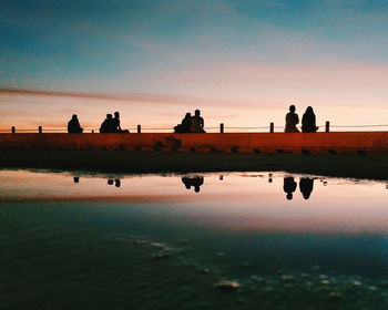 Silhouette people reflecting on water at beach against sky during sunset