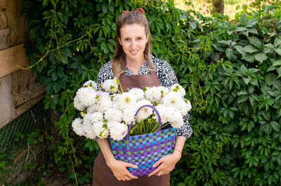 A cute girl in an apron with a basket of white dahlias flowers stands near the vineyard and smiles