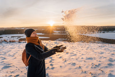 A woman in winter in warm clothes throws her palms and snow against the background of sunset.