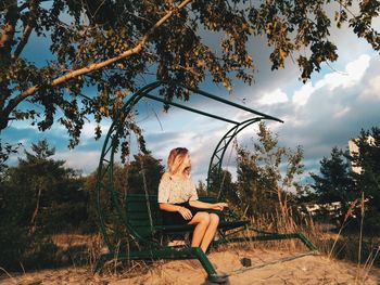 Woman sitting on swing at park against cloudy sky
