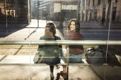 Smiling woman looking over shoulder while sitting with female friend at tram stop seen through glass