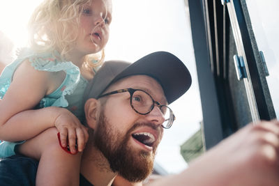 Low angle view of father talking while carrying daughter on shoulders during sunny day