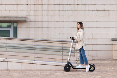 Side view of young woman riding sustainable electric scooter in the city in front of white building