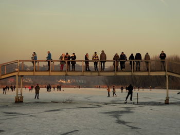 People on bridge over frozen lake against sky during sunset
