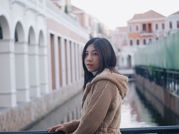 Portrait of young woman standing on footbridge against buildings in city