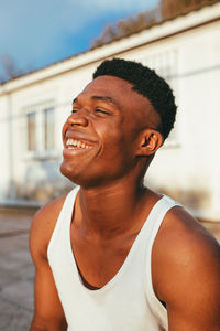 Happy african american male in undershirt with modern haircut looking forward against building in sunlight