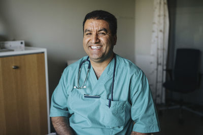 Portrait of cheerful mature male doctor in scrubs sitting in clinic