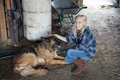 A girl in cowboy-style clothes is sitting in a stable and stroking german shepherd