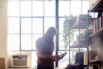 Man reading book while standing by window at home