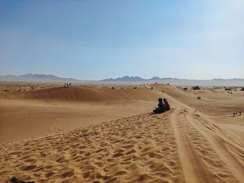 Rear view of man sitting on sand at desert against clear sky