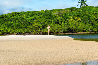 A woman is on the sand next to the red water river against the green forest in the background. 