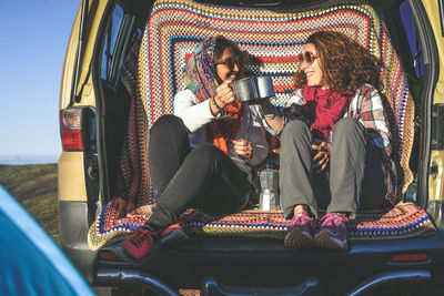 Smiling friends toasting coffee mugs in car trunk