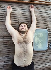 Portrait of shirtless man with arms raised against brick wall