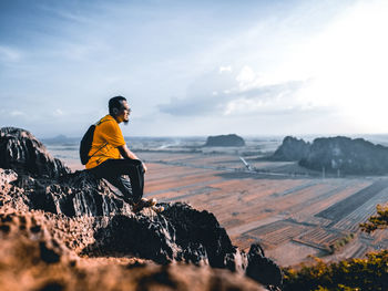 Side view of man sitting on rock against sky