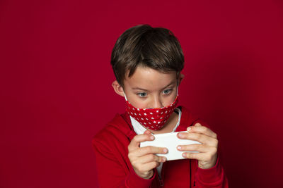 Boy wearing mask using mobile phone against red background