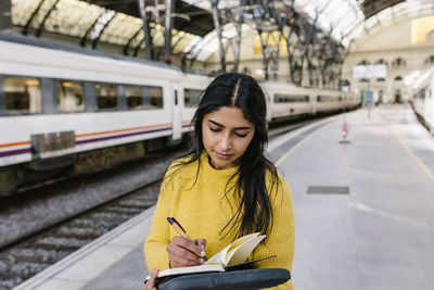 Woman writing in book while standing on railway platform