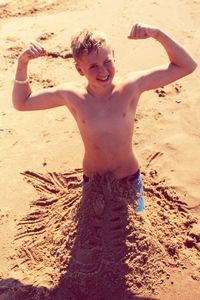 High angle portrait of smiling boy flexing muscle while standing in sand at beach