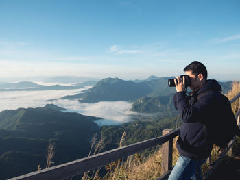 Side view of man photographing while standing on mountain against sky