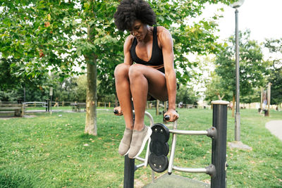 Low angle of positive young curly haired black sportswoman doing push ups on parallel bars in summer park