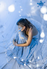 High angle view of woman with make-up sitting by illuminated lighting equipment on bed