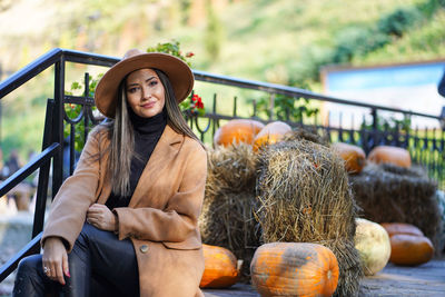 Portrait of smiling young woman wearing hat sitting by pumpkins