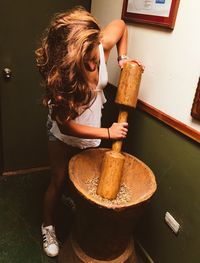 Young woman grinding ingredients using large mortar and pestle against wall