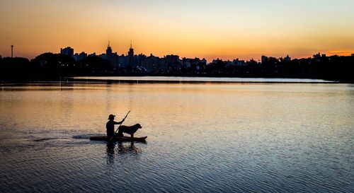 Silhouette man paddleboarding with dog on lake against city