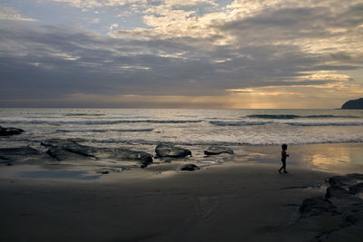 Side view of boy walking at beach against sky during sunset