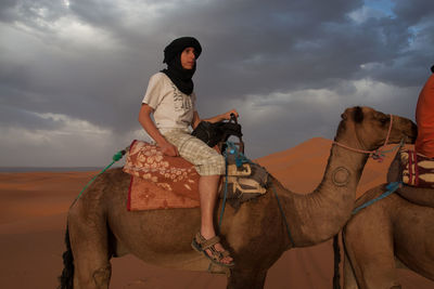 Side view of young man on camel at desert against cloudy sky