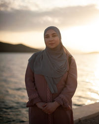 Portrait of young woman standing against sea during sunset