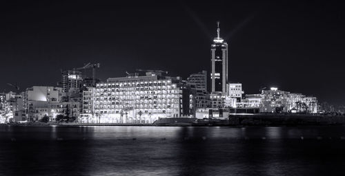 Illuminated buildings by sea against sky at night