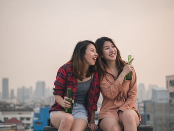 Cheerful friends holding bottles looking away against sky in city