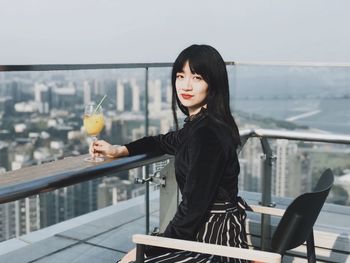 Portrait of young woman with drink sitting in balcony against cityscape