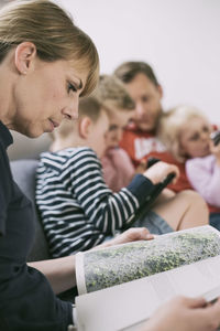 Side view of woman reading magazine with family using technologies in background