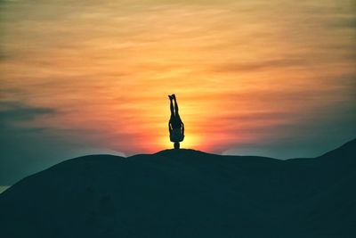 Silhouette man practicing headstand on mountain against sky during sunset
