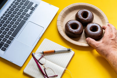 Male hand taking a chocolate donut in the workplace, next to a laptop on yellow bacground.