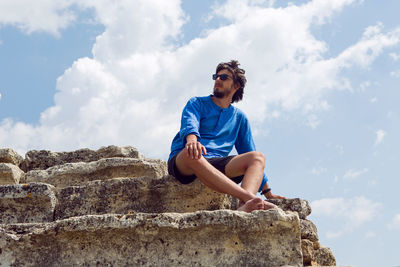 Man in a blue linen shirt and shorts sits on an old stone staircase  colosseum in pamukkale turkey