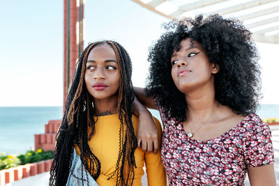 Thoughtful black woman with curly hair leaning on shoulder of african american female friend with braids while standing on embankment and looking away