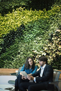 Businessman and woman sitting in front of green plant wall, using digital tablet