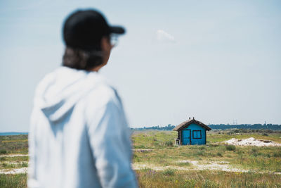 Rear view of man standing on field and looking at blue house against clear sky in summer