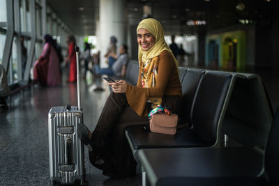 Portrait of woman holding mobile phone while sitting by suitcase at airport