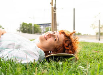 Pleased female with freckles in casual wear lying looking at camera on skateboard on grassy lawn on summer day