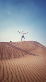 Mid distance view of man with arms outstretched standing on sand dune against blue sky