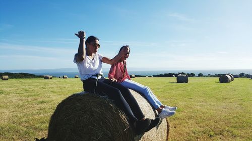 Female friends sitting on hay bale against sky during sunny day