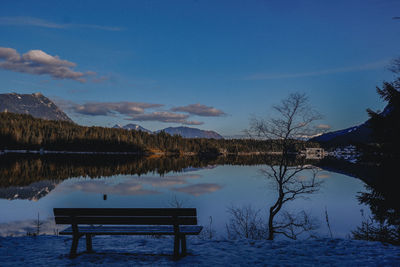Bench by lake against blue sky during winter