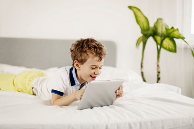 Curly-haired boy is lying on the bed in the bedroom and playing computer games on a tablet