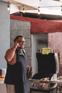 Smiling ethnic male talking on smartphone while standing with bottle of beer at barbecue cooker with open lid and preparing food