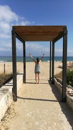 Rear view of woman with arms raised standing on beach against sky