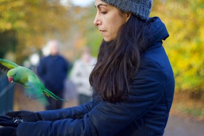 Side view of young woman holding autumn leaf against trees